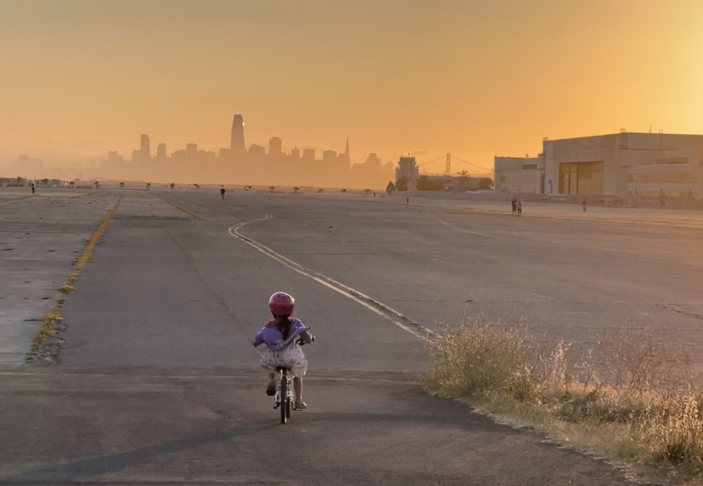 A child on a bike rides towards the San Francisco skyline against an orange sunset.