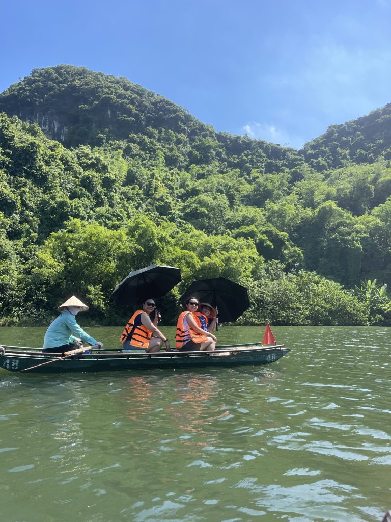 passengers sit under umbrellas while a rower wearing a teal long-sleeved shirt and a Vietnamese conical hat steers the boat.