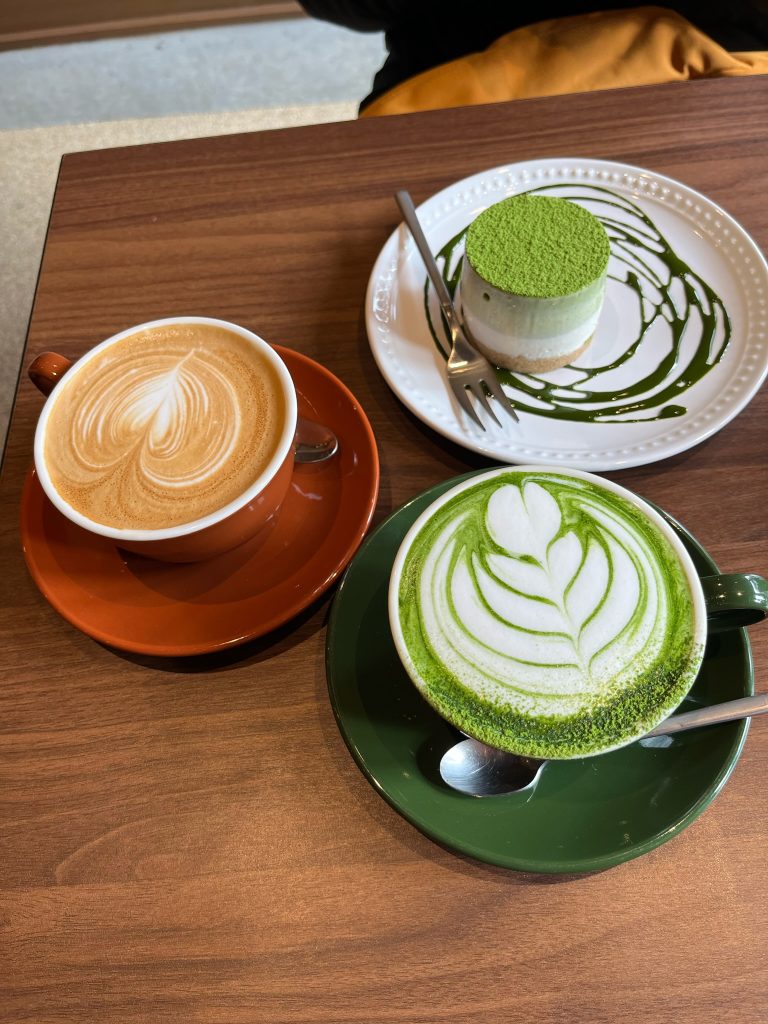 A mug each of matcha and a latte, as well as a matcha cheesecake plated with chocolate swirl.