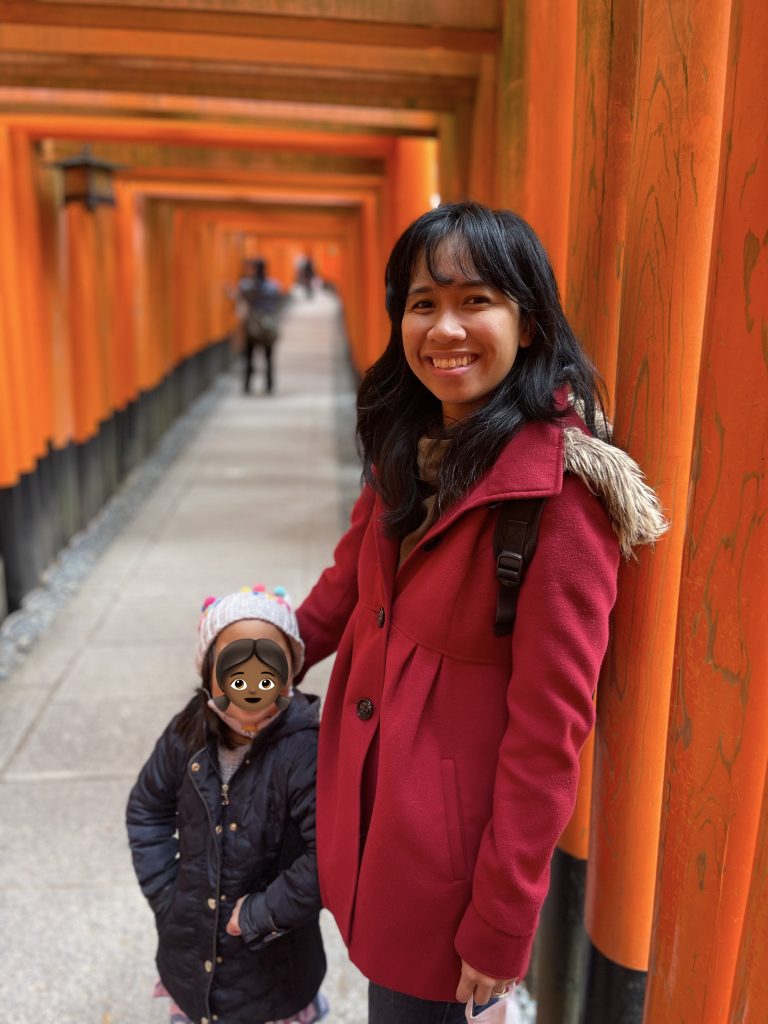 Jelly poses at the camera in a red coat with her arm around her kid, wearing a blue coat. They are surrounded by orange pillars.