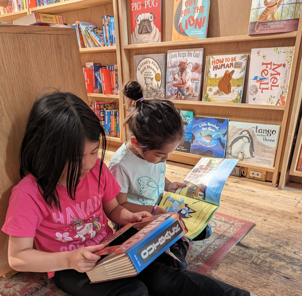 Two young girls read books in front of a bookshelf full of children's books.