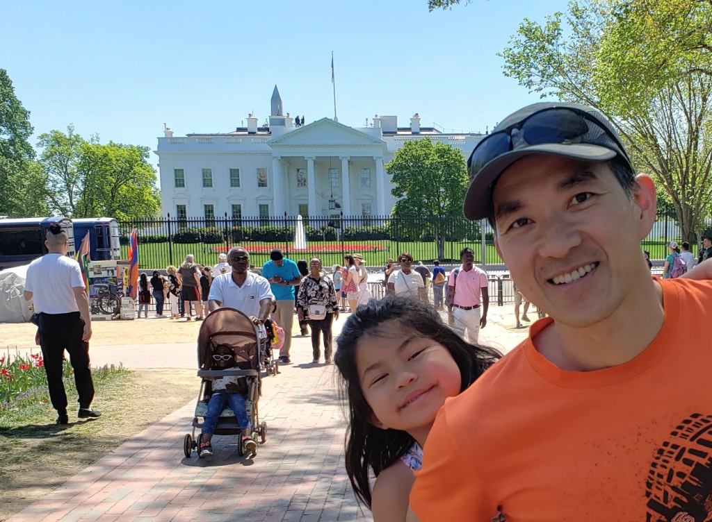 A dad in an orange tshirt and grey hat smiles at the camera as his young daughter peeks out to the camera from over his shoulder in front of a crowd in front of the White House.