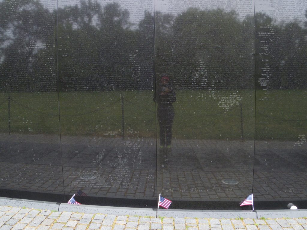 A black reflective wall with hundreds of names inscribed reflects back an individual wearing black amidst a grassy, tree-lined background.