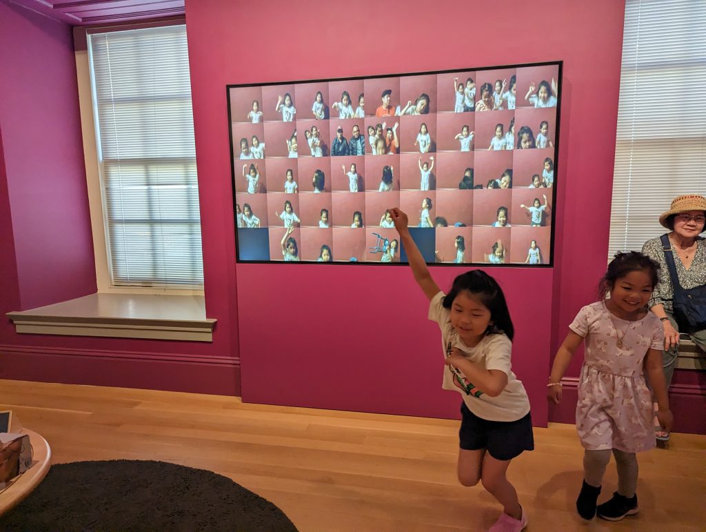 A child in a white shirt lands a jump in front of a pink wall with a screen full of > hanging