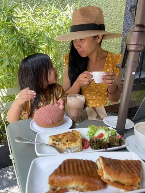Against a green leafy background, Jelly in a yellow dress, is seated next to her kid ready to eat their brunch spread!